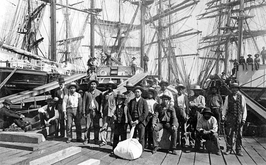 Nineteenth century dock workers posed on a wharf in front of tall sailing ships