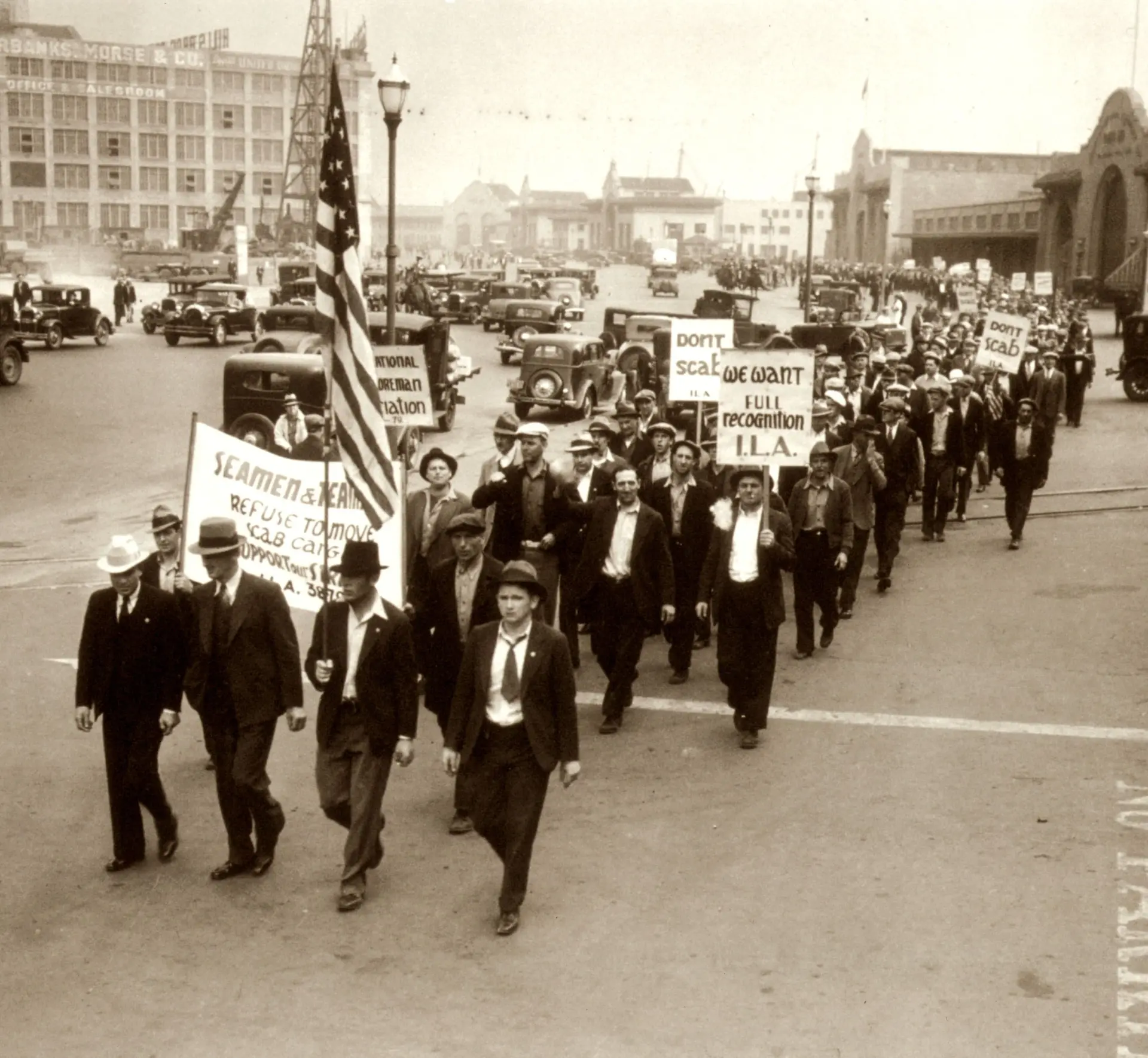 Longshore workers marching down a waterfront street carrying union picket signs and an American flag